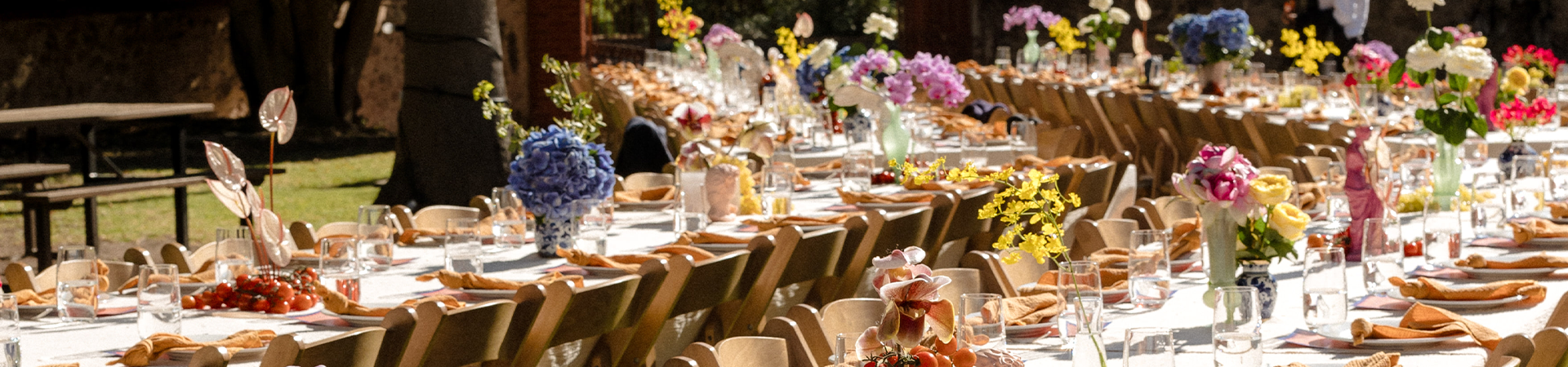 Two long curved tables set with white tablecloths, topped with multi-coloured floral arrangements, glassware and crockery. Tables are set with timber chairs.