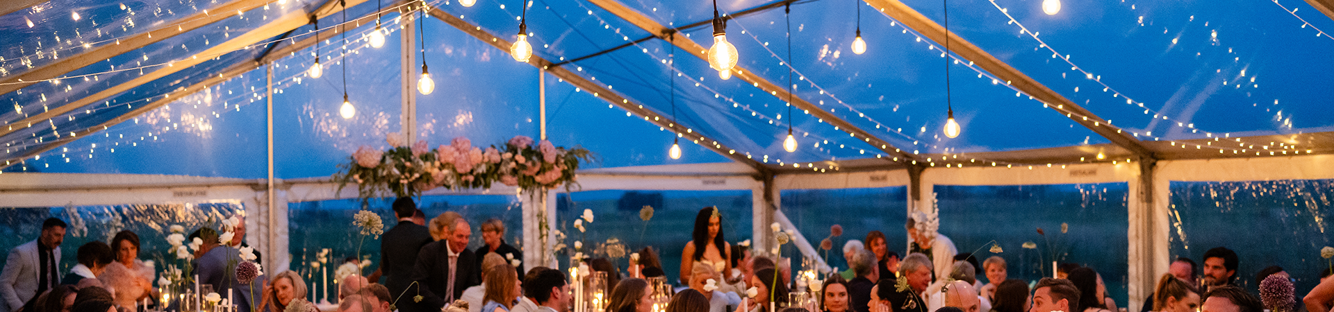 A clear marquee with festoons and fairy lights in the evening, with people below enjoying a wedding dinner